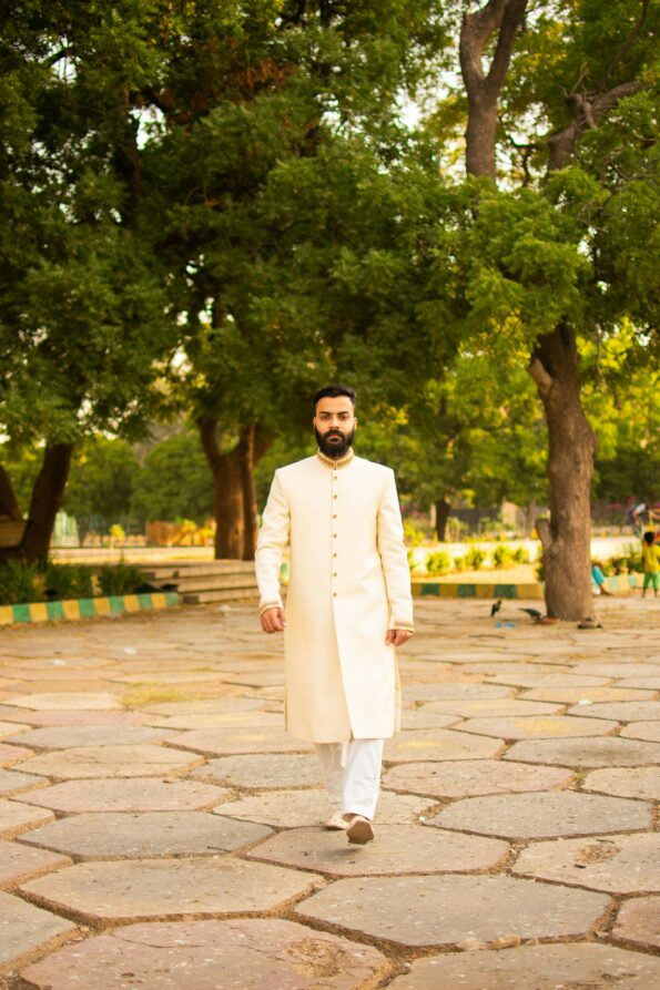 Indian man in traditional attire walking through a sunlit park surrounded by lush trees.